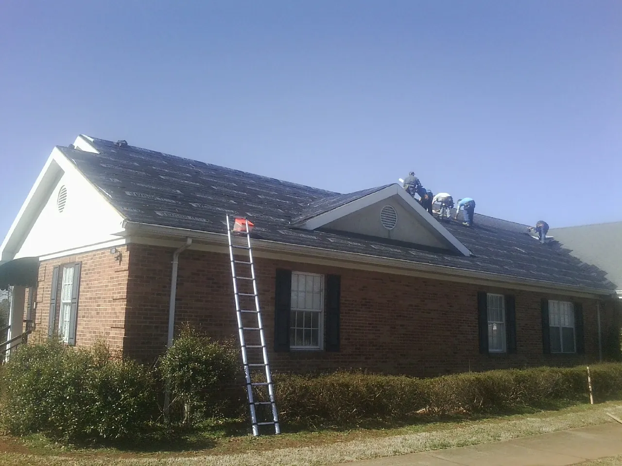 Crew tearing shingles off a brick ranch roof with a ladder set — Metal Roof Repair work by our crew in Cowen
