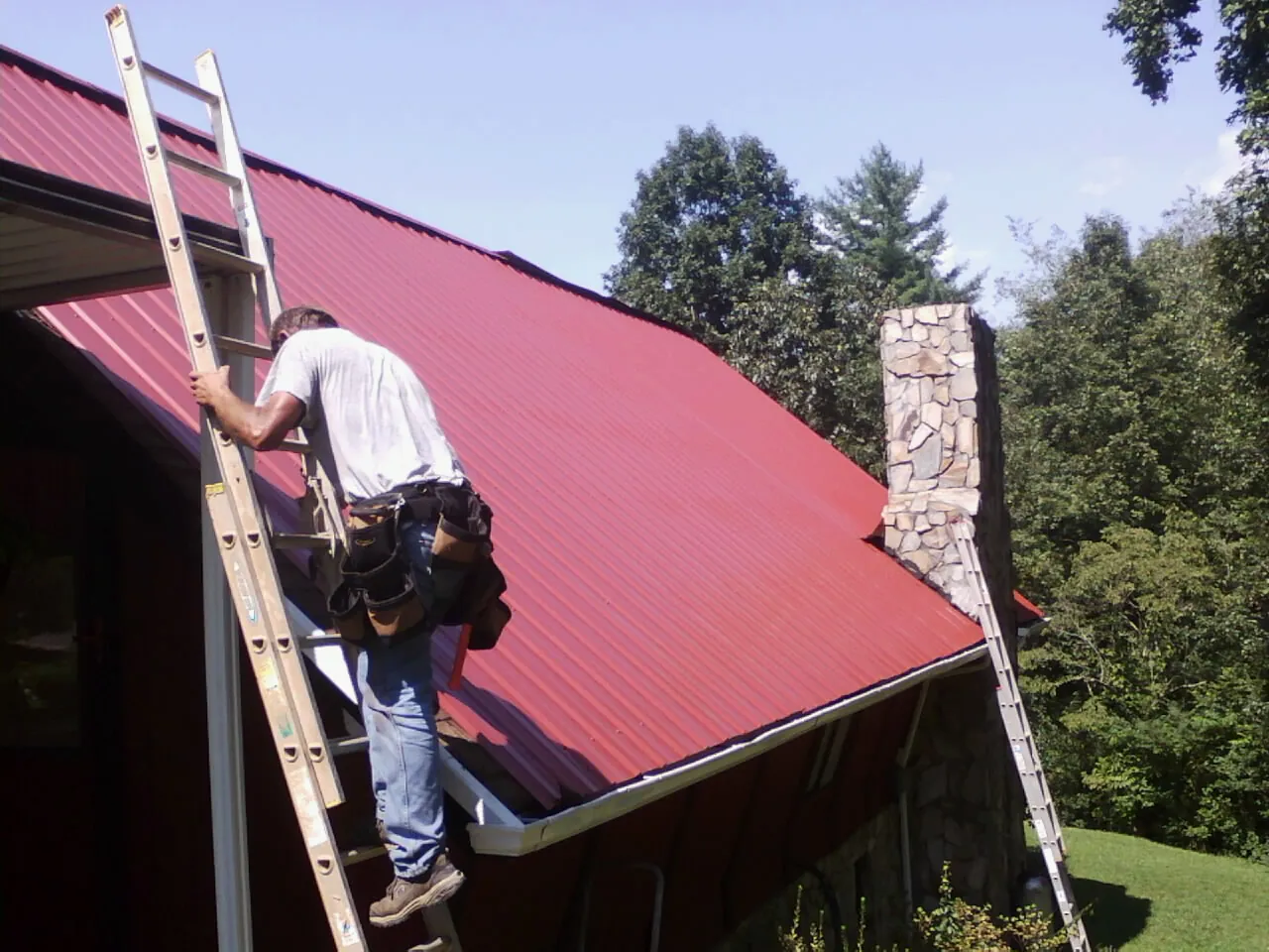 Roofer on a ladder beside a red metal roof and stone chimney — Soffit & Fascia Repair by Roofing Brothers in Cowen
